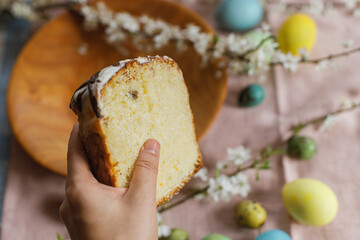 Hand holding half of baked easter cake on background of natural dyed easter eggs with spring flowers on wooden plate on rustic table.  Traditional Easter food. Happy Easter!