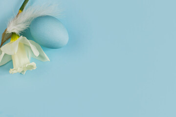 Happy Easter! Easter egg and flower on blue background flat lay. Minimal Easter still life. Modern spring banner or greeting card, space for text. Natural blue egg, feather and daffodil