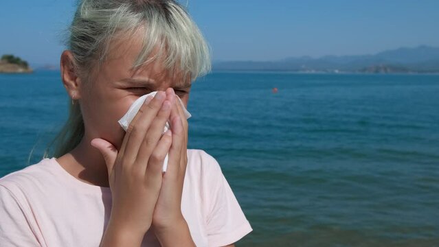 Allergic rhinitis. A young girl sneezes outdoors on the seashore.