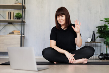 young woman sitting on yoga mat and doing yoga with  online tutorial on laptop