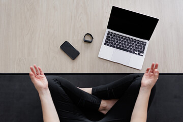 sport and technology concept - top view of young woman sitting in lotus pose on yoga mat, laptop and smartphone with blank screens and fitness tracker