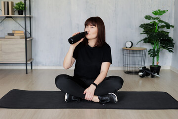 tired young woman sitting on yoga mat and drinking water after training