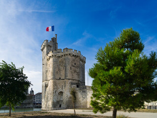 Medieval tower with the French flag at the entrance of the port of La Rochelle with blue sky and light white clouds.