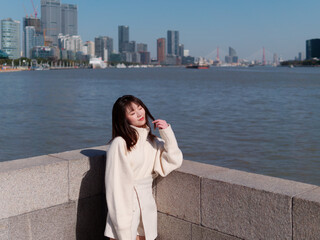 Beautiful young woman with black long hair in white skirt posing eyes closed with blur Shanghai Bund landmark buildings background in sunny day. Emotions, people, beauty, travel and lifestyle concept.