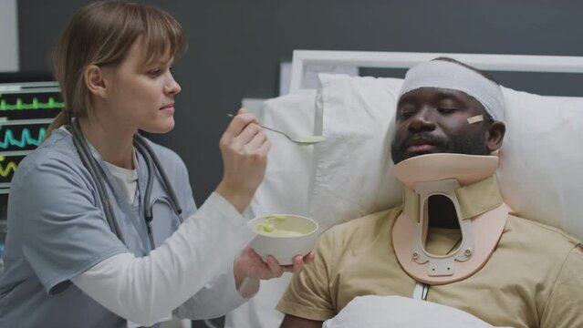 Professional nurse spoon feeding injured African American man with bandage on head and neck brace in military hospital