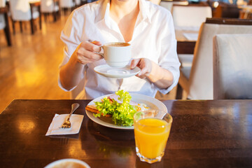 No face a woman drinks coffee at breakfast in the hotel restaurant. Salad, eggs, orange juice,...
