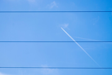 A passenger plane and its trail of white smoke behind some high voltage wires against the blue sky.