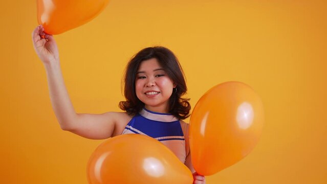 joyful cheerleader in white-blue uniform holding orange balloons, medium closeup, studio shot, orange background. High quality 4k footage