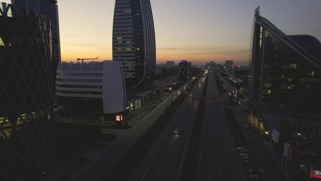 Drone footage of the bustling streets of Sofia's business district at dusk, as the city's skyline is bathed in the warm glow of the setting sun. The camera follows the flow of traffic, weaving through