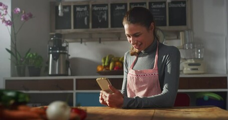 Portrait of a Woman in the Kitchen, Wearing an Apron, Using a Smartphone and Laughing. Female Chef Scrolling, Checking Social Media, Choosing Recipe, Shopping Online. Housewife Staying Connected
