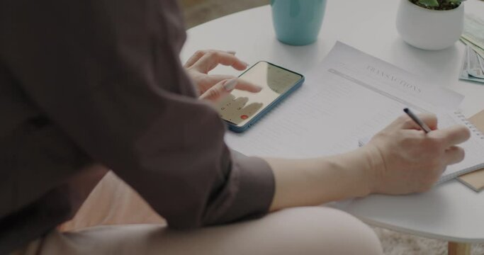Close-up of female hands using calculator in smartphone and writing financial information on paper counting money at home. People and accounting concept.