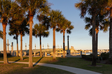 Golden hour view of the 1927 double-leaf bascule Bridge of Lions connecting downtown to Anastasia Island across Matanzas Bay and harbour piers through palm trees, St. Augustine, Florida