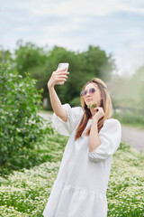 Fototapeta premium Young millennial woman making selfie in chamomile field, using technology. Blogging lifestyle. Relaxing and leisure