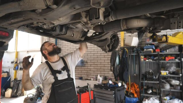 Professional bearded auto mechanic working underneath a lifting vehicle at service or garage. Repairman repairing car with special tool at workshop. Concept of automobile maintenance. Slow motion
