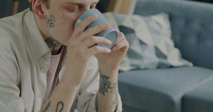 Portrait of creative young man in trendy outfit drinking tea from mug and watching TV indoors at home. Beverage and leisure time activity concept.