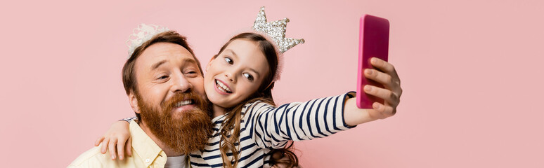 Positive child hugging father with crown headband while taking selfie isolated on pink, banner.