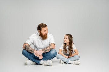 Fototapeta na wymiar Smiling girl talking to tattooed father while sitting on grey background.