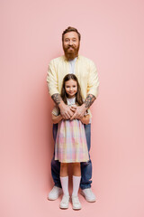 Full length of smiling tattooed man hugging daughter on pink background.