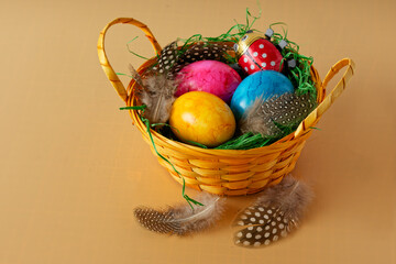 Several colored Easter eggs in a yellow woven basket on a yellow background. Decorated with some chicken feathers.
