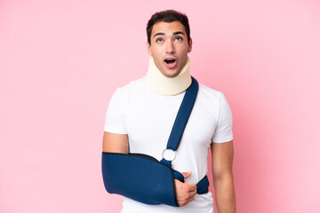 Young caucasian man with broken arm and wearing a sling isolated on pink background looking up and...