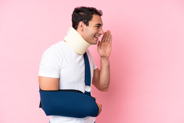 Young caucasian man with broken arm and wearing a sling isolated on pink background shouting with...