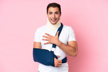 Young caucasian man with broken arm and wearing a sling isolated on pink background surprised and...