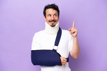Young caucasian man wearing a sling and neck brace isolated on purple background pointing up a...