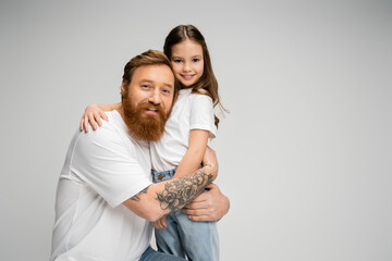 Smiling tattooed dad hugging happy daughter isolated on grey.
