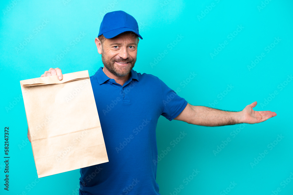 Wall mural Middle age man taking a bag of takeaway food isolated on blue background extending hands to the side for inviting to come