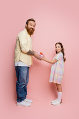 Positive girl giving gift to bearded dad and looking at camera on pink background.