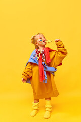 Full-length photo of little girl wearing big clothes, smiling with lollipop to camera over yellow background. Concept of fashion, ad, children's products, childhood, child model