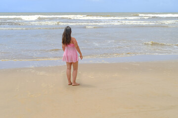 woman with long hair in pink swimsuit stands barefoot on sandy seashore, enjoys sea breeze, takes sunbaths, blue waves to horizon, concept travel, vacation in tropical countries, summer sun protection