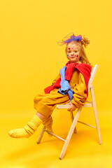 Photo of sweet little girl wearing bow, sitting on chair and dangling feet over yellow background
