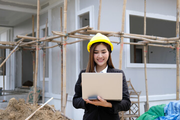 Portrait of a Happy Young Beautiful Female Engineer Wearing yellow Hard Hat, Using Laptop Computer in Office at Car Assembly Plant. Industrial Specialist Working on Vehicle Design in Modern Facility.