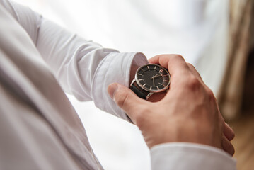A man in a white shirt puts an expensive watch on his hand and holds the dial with his fingers checking the time.