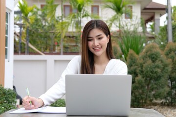 Young woman works on laptop computer while sitting relaxed on chair on background of green bushes at backyard. Concept of remote work at cozy atmosphere