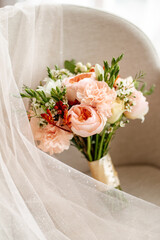 A bouquet of fresh pins and other flowers, tied with a beige ribbon, stands on a neatly laid out, delicate, cream-colored bridal veil with a splash of sequins.