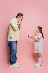 Side view of shocked man looking at preteen daughter with gift on pink background.