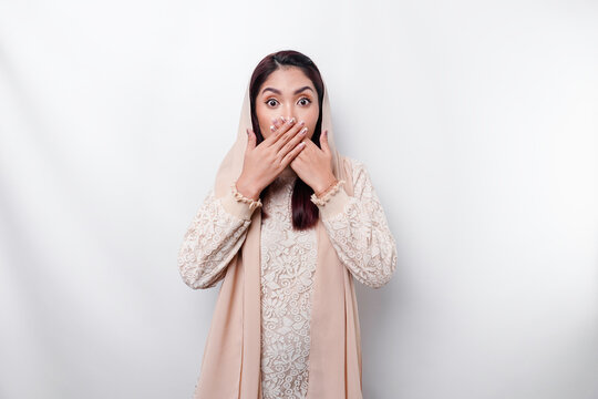 Oops! Surprised young Asian Muslim woman covering mouth with hands and staring at camera while standing against white background