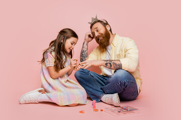 Cheerful girl applying nail polish on hand of tattooed dad with crown headband on pink background.