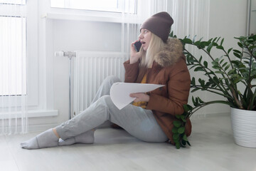 A woman warmly dressed in a brown jacket and hat is sitting near the heater with bills in her hands and shouting on the phone. The concept of a crisis and a large set for utilities.