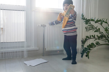 The child is warmly dressed in a sweater and a hat, standing and holding on to the radiator with money and bills. The concept of the economic crisis and the lack of heat and heating in homes.