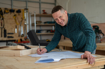 Portrait of a carpenter in a plaid shirt draws a workshop blueprint. 