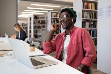 Happy excited African American man student sits in library at table with laptop and laughing...