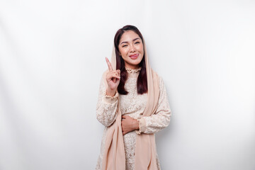 Starving Asian Muslim woman wearing a headscarf, pointing up at the copy space above her, isolated by a white background