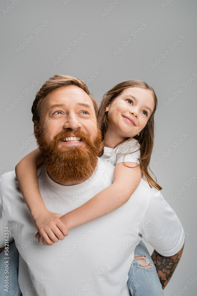 Wall mural Smiling girl piggybacking on bearded parent and looking away isolated on grey.