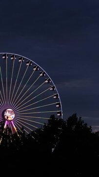 Close-up of a neon coloured ferris wheel rotating against the dark blue clouds. Beer festival, carnival, amusement or theme park after sunset, entertainment and fun at night.