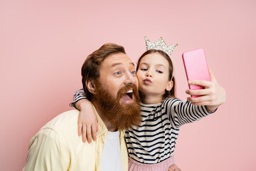 Preteen girl with crown headband pouting lips and taking selfie with bearded dad isolated on pink.