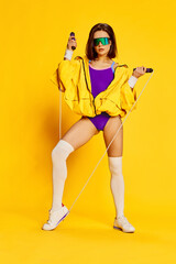Fototapeta na wymiar Fitness trainer. Full-length image of stylish young woman in bright sportswear and sunglasses posing with jumping rope against yellow studio background. Concept of sport, healthy lifestyle, beauty. Ad