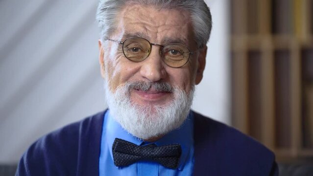Portrait of senior man grandfather teacher with a gray beard wearing glasses with a bow tie looking at the camera smiling tilting his head to the side